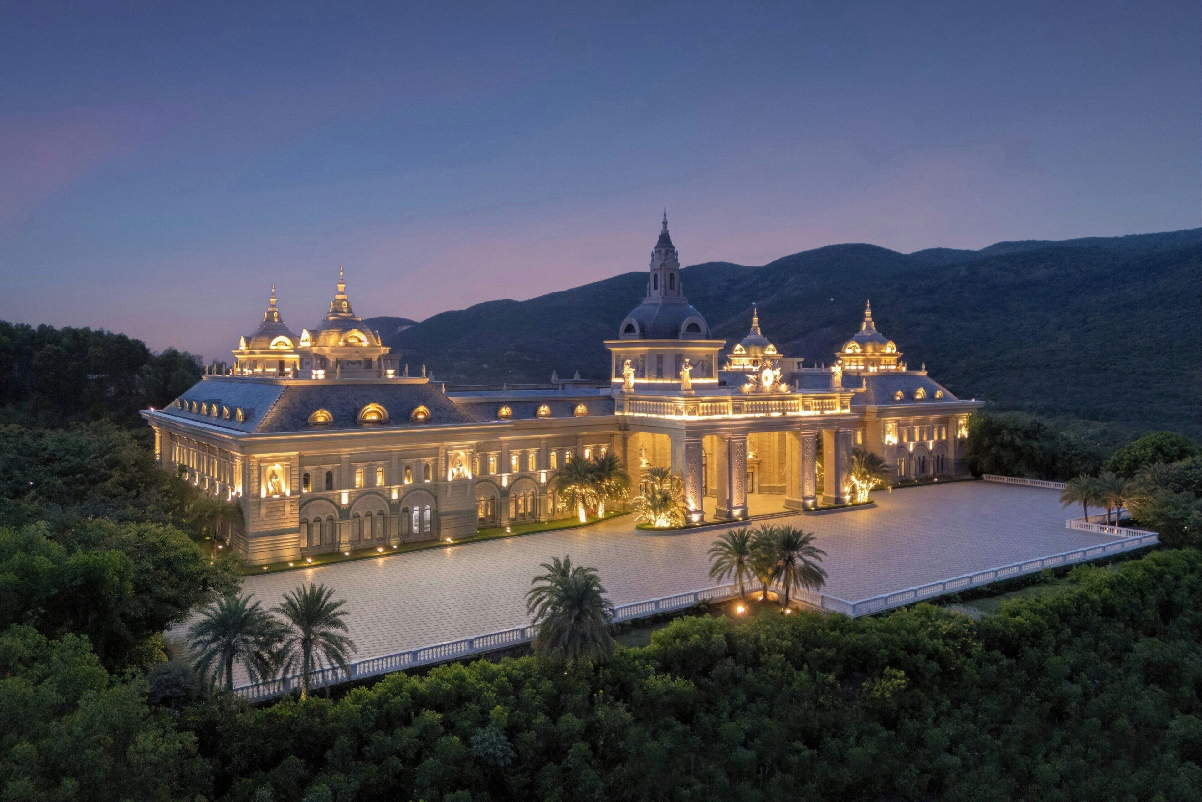 A large, illuminated building with multiple domes and a central tower, surrounded by lush greenery and a paved courtyard.