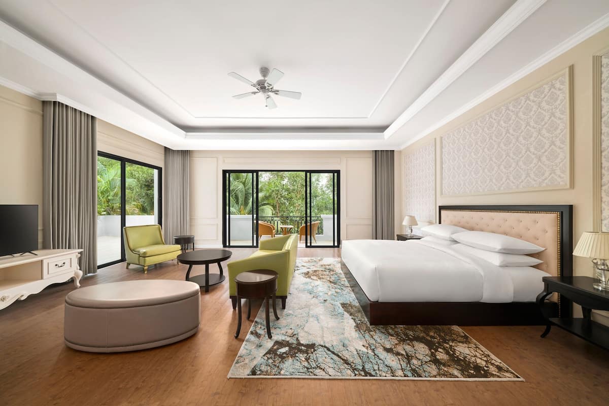 Habitación de hotel con una cama grande, televisión, sofá, mesa de centro y balcón con mobiliario exterior.