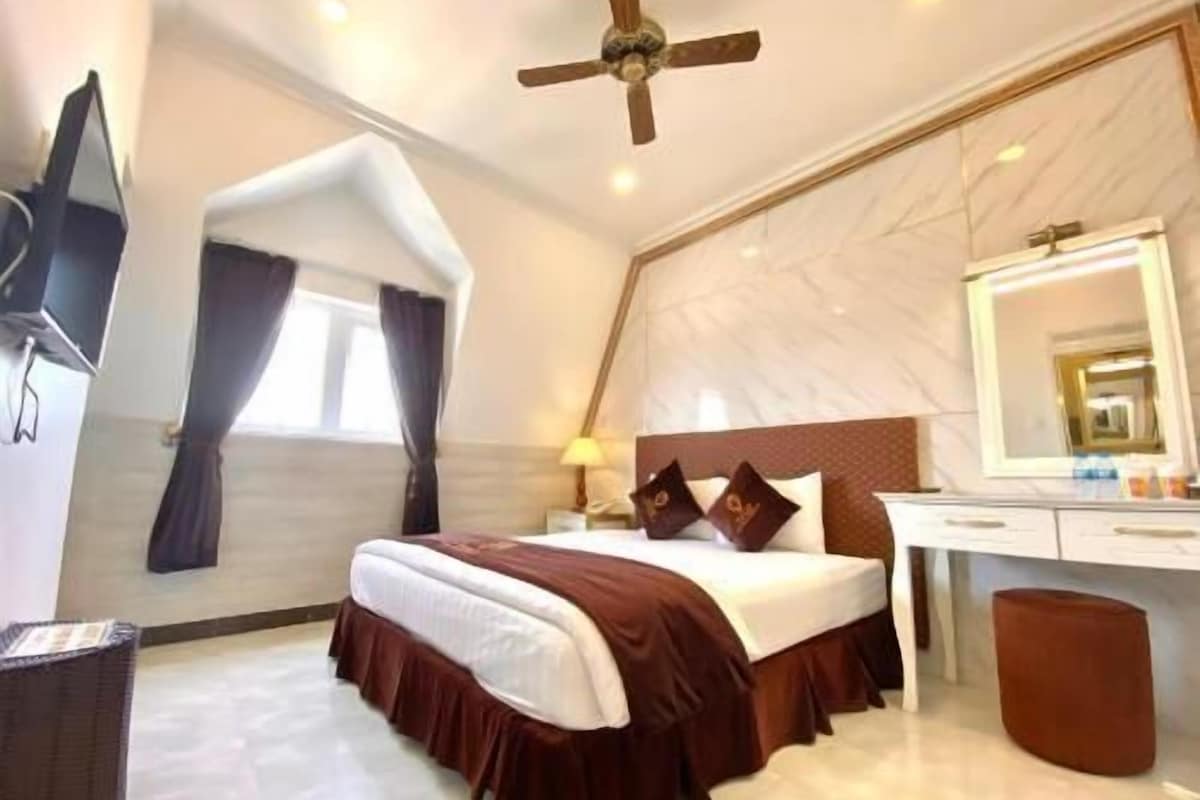 A hotel room with a bed, a desk, a mirror, a ceiling fan, and a window with curtains.