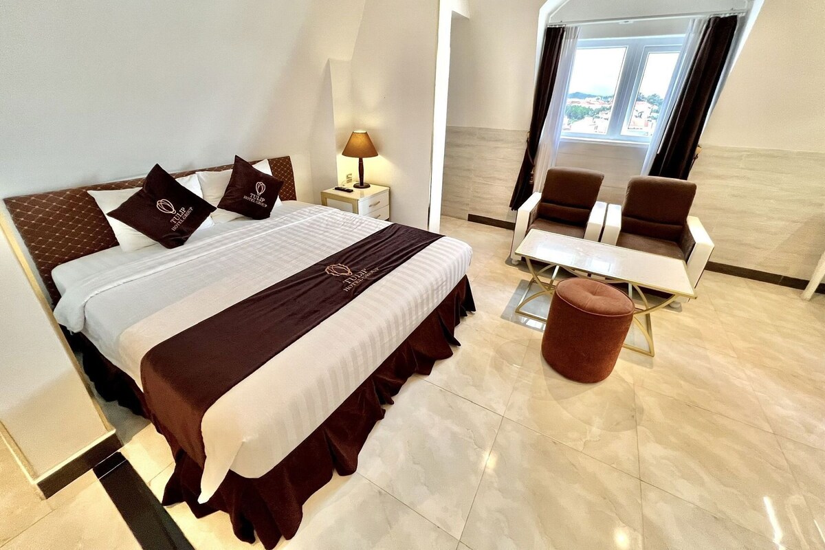 A hotel room with a large bed, a desk, two chairs, a lamp, and a view of the city.