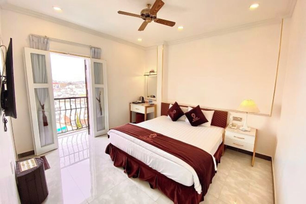 A hotel room with a large bed, a ceiling fan, a bedside table with a lamp, and a view through a sliding door to the outside.