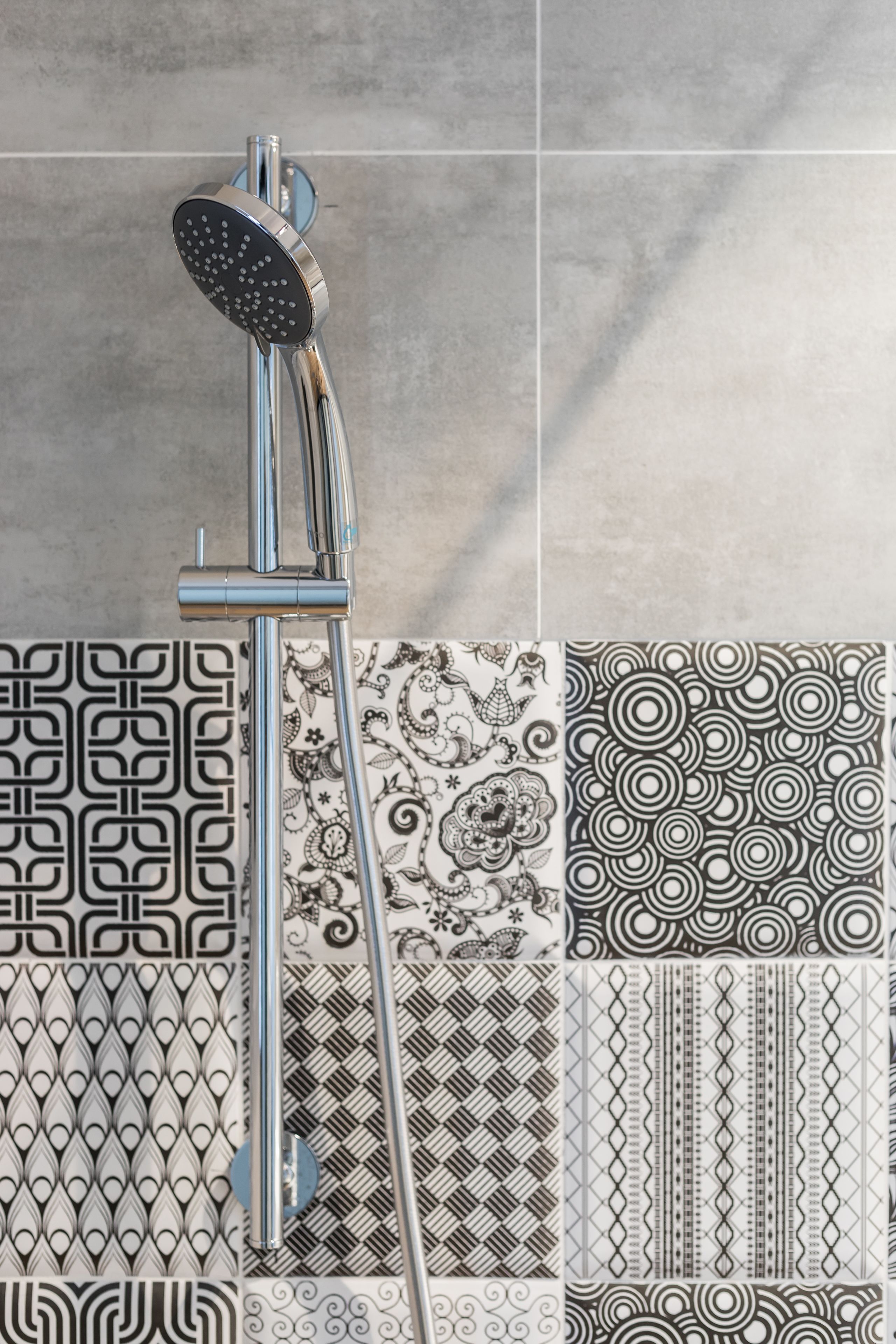 A showerhead mounted on a chrome fixture with a tiled wall featuring various geometric and floral patterns.