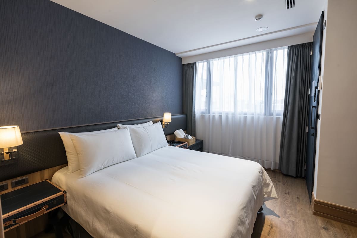 A hotel room with a large bed, a nightstand with a lamp, a suitcase, and a window with curtains.