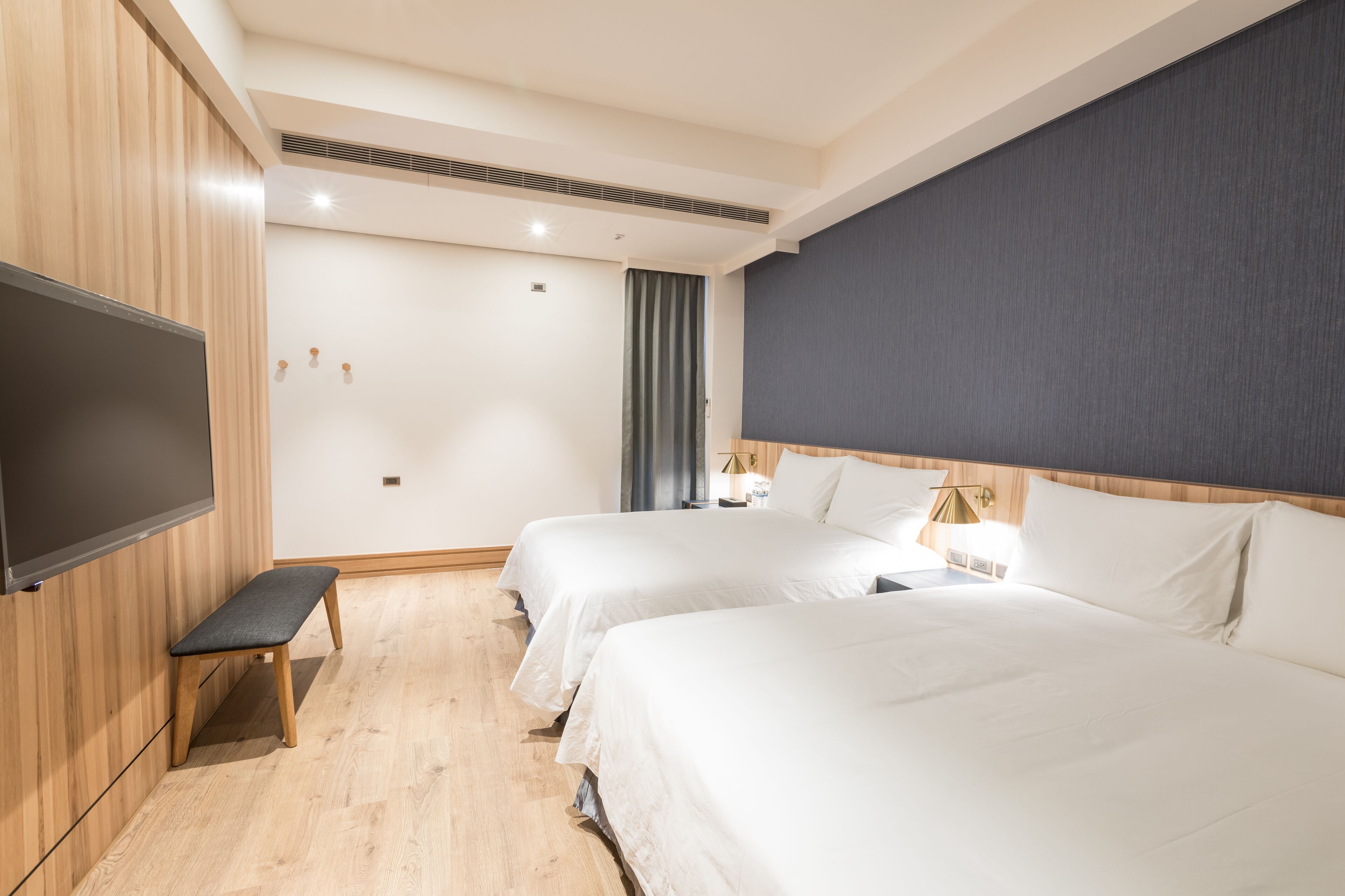 A hotel room with two beds, a flat-screen TV, a bench, and a nightstand with a lamp.