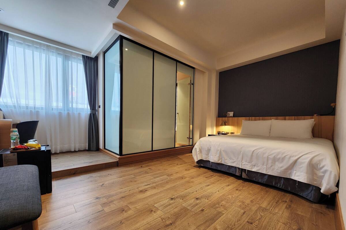 A hotel room with a bed, a desk, a chair, a wardrobe, and a large window with curtains.