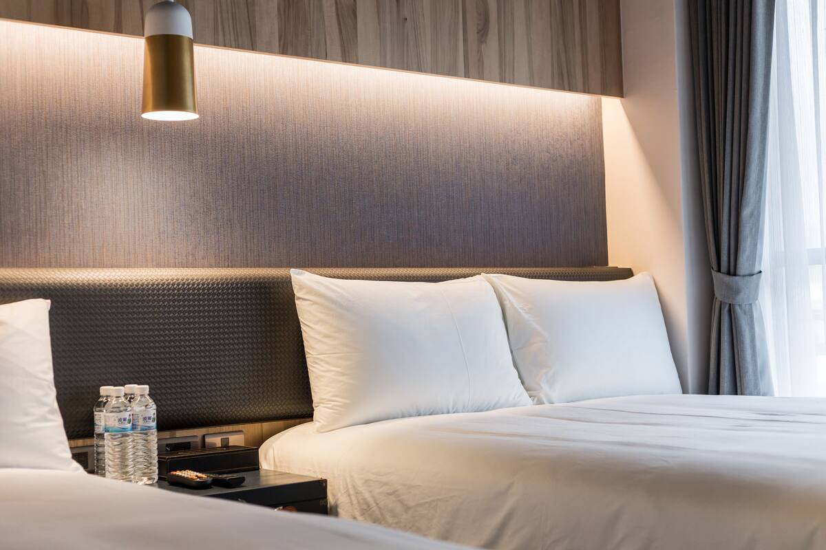 A hotel room with a large bed, a bedside table with water bottles, a remote control, and a lamp.