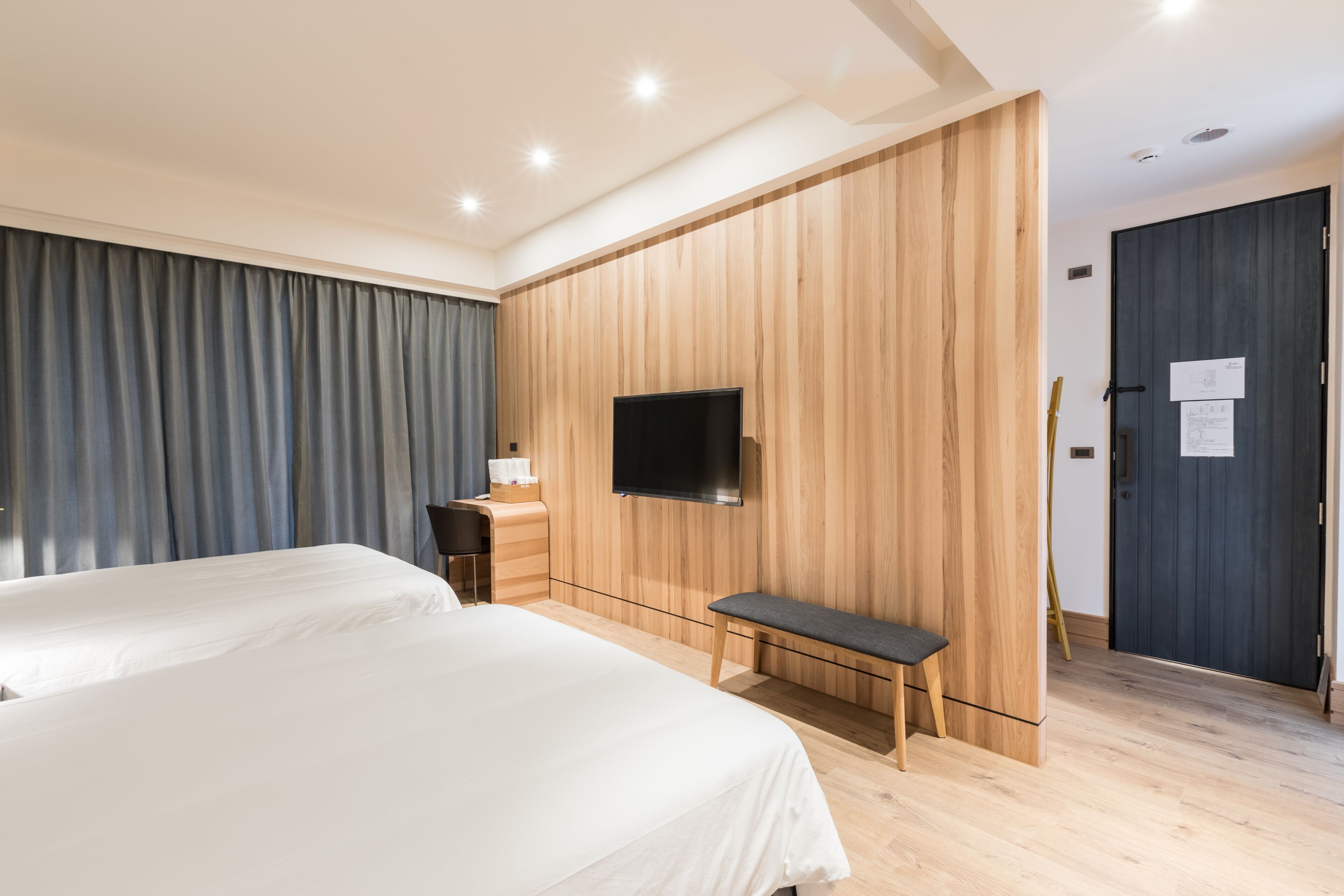 A modern hotel room with a large bed, a desk with a chair, a flat-screen TV mounted on a wooden wall, and a bench.