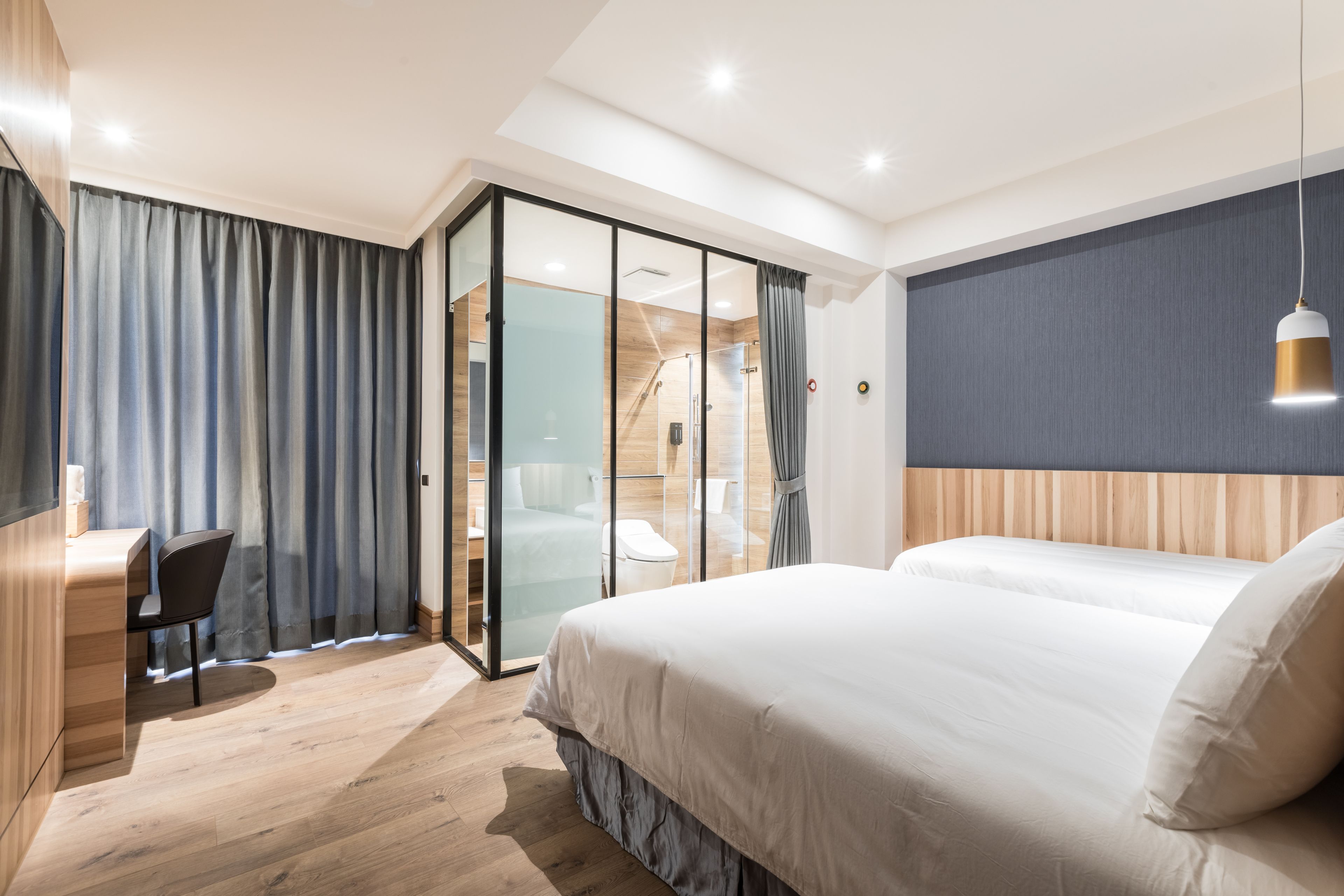 A modern hotel room with a large bed, a desk, and a glass partition separating the room from a shower area.