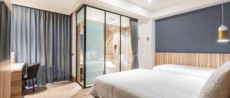 A modern hotel room with a large bed, a desk, and a glass partition separating the room from a shower area.