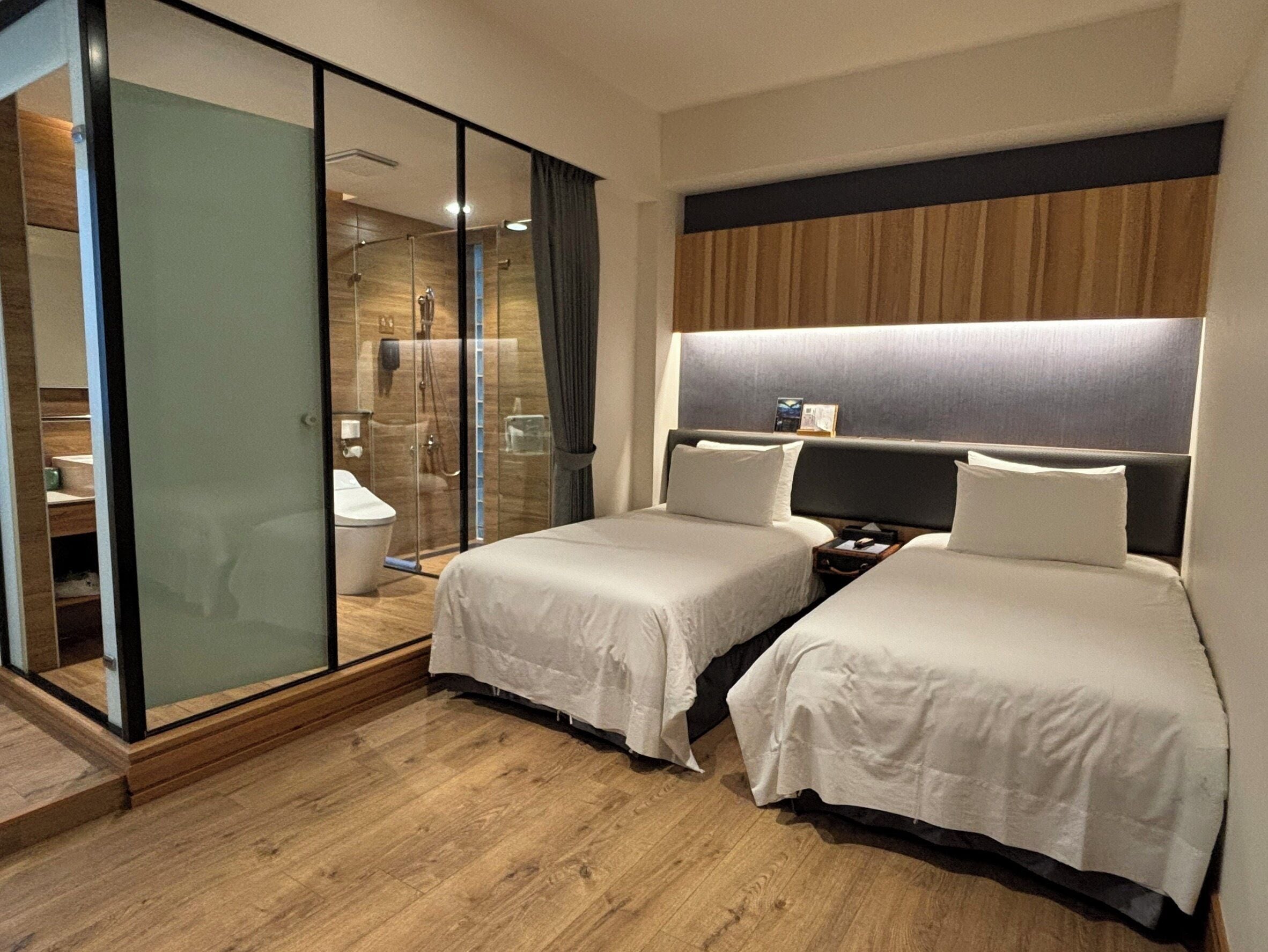 A hotel room with two beds, a wooden floor, a glass shower enclosure, and a modern headboard.