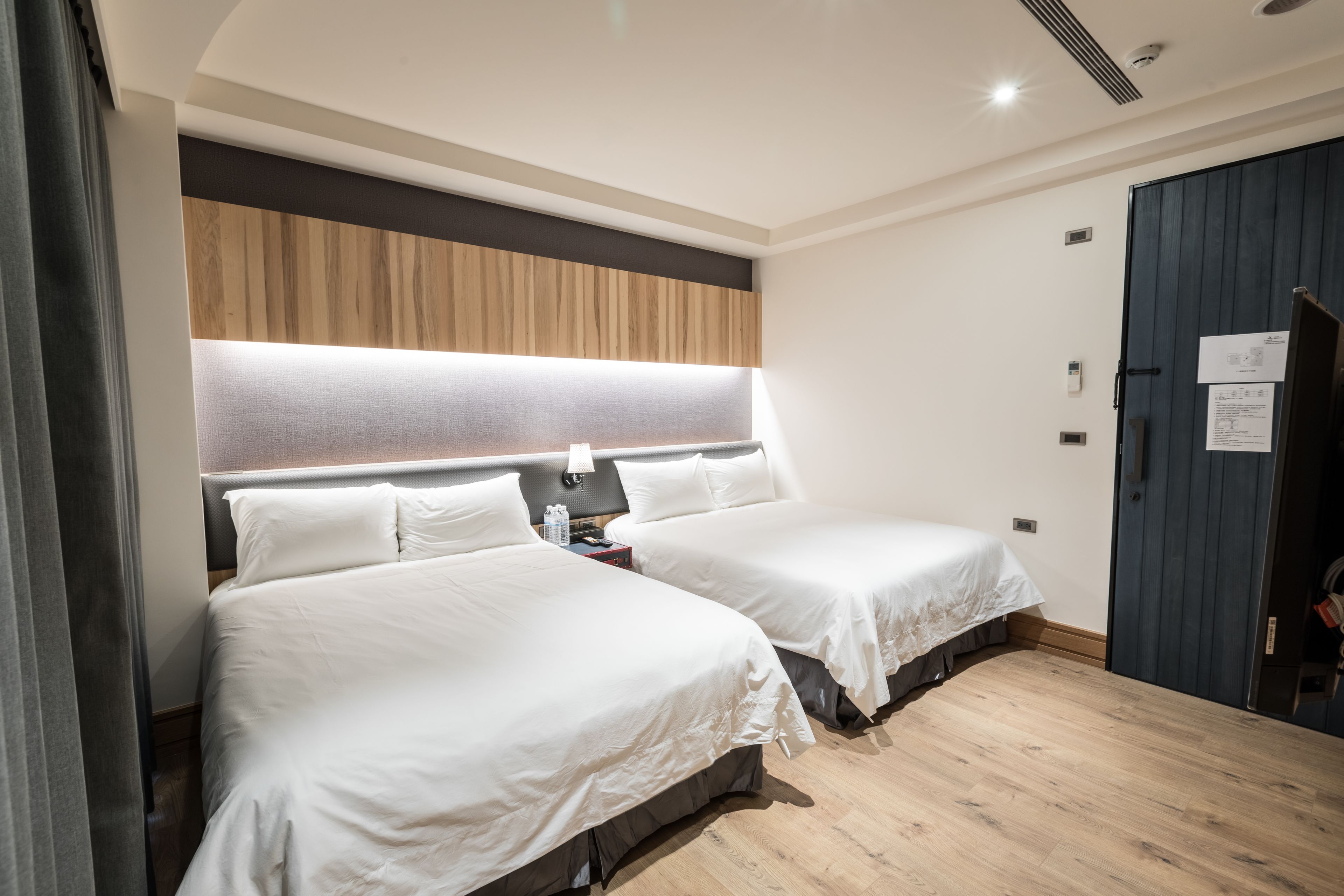 A hotel room with two beds, a wooden headboard, a bedside table with a lamp, and a door with a notice.