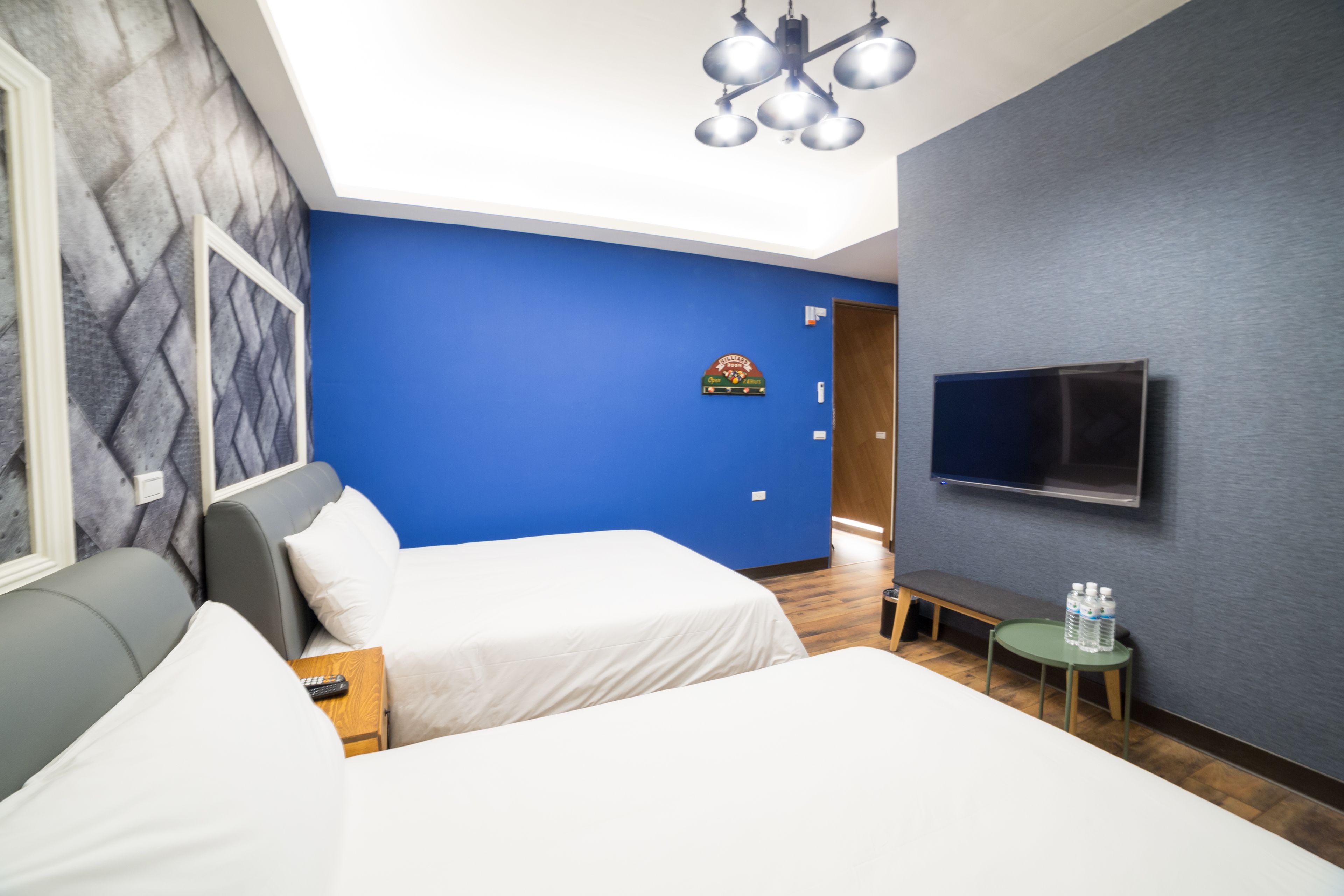 A hotel room with two beds, a TV mounted on the wall, a small table with water bottles, and a bench.