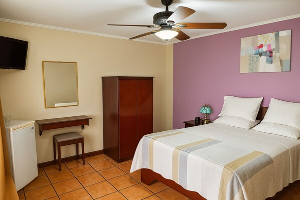 A hotel room with a bed, a desk, a wardrobe, a ceiling fan, a television, and a painting on the wall.