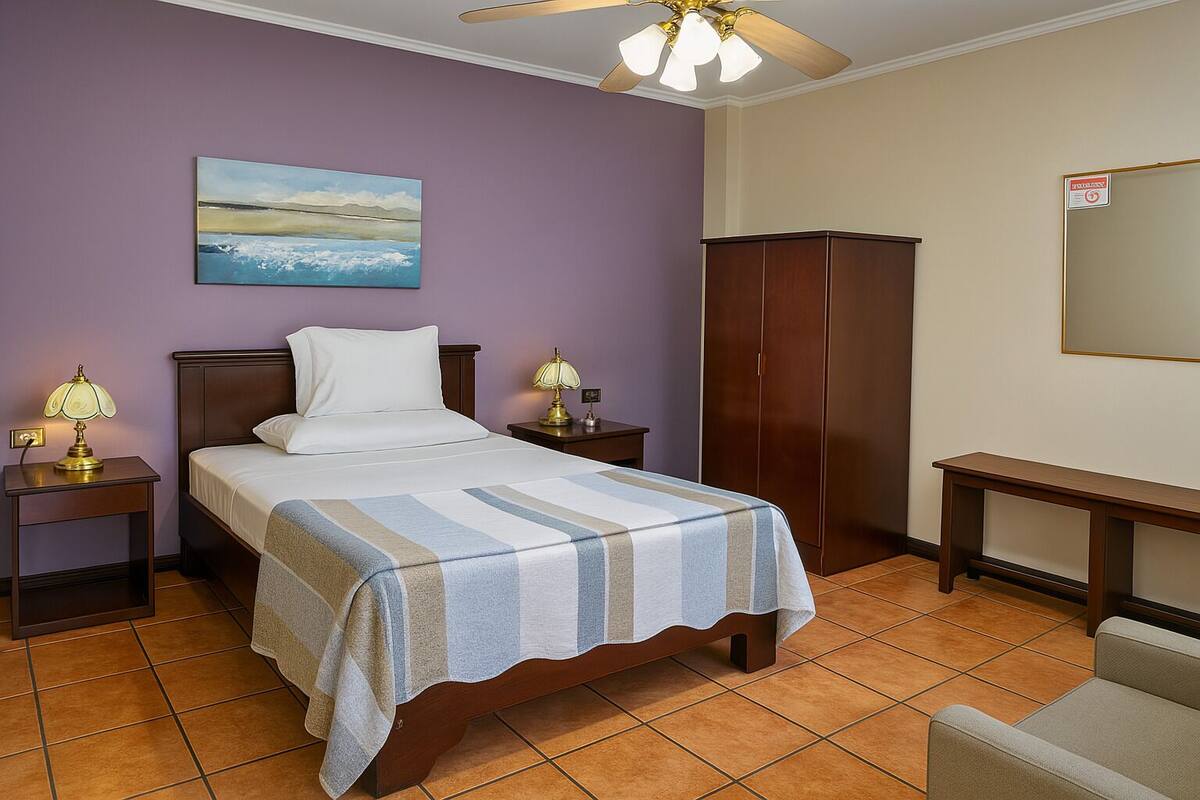 A hotel room with a bed, two bedside tables with lamps, a wardrobe, a bench, and a painting on the wall.
