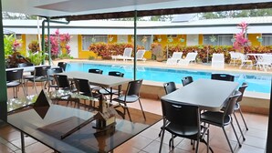 Een zwembadterras met ligbedden en tafels, met een helder uitzicht op het zwembad, en een geel gebouw op de achtergrond.