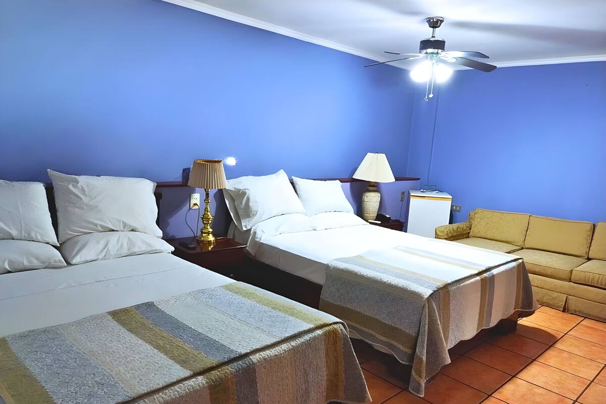 A hotel room with two beds, a ceiling fan, a small table with a lamp, and a sofa.