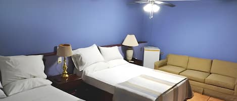 A hotel room with two beds, a sofa, a ceiling fan, and a nightstand with a lamp.