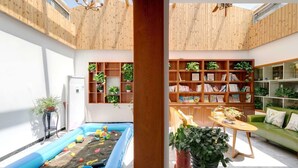 Un espace de vie moderne avec une piscine gonflable bleue, des étagères en bois ornées de plantes, un canapé vert et un plafond en bois.