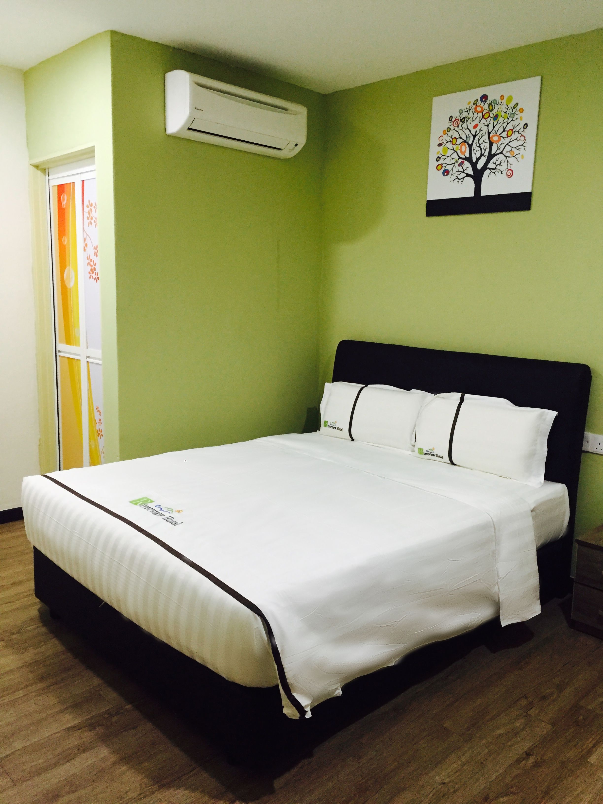 A neatly made bed in a room with a green wall, a painting of a tree, and an air conditioning unit above it.