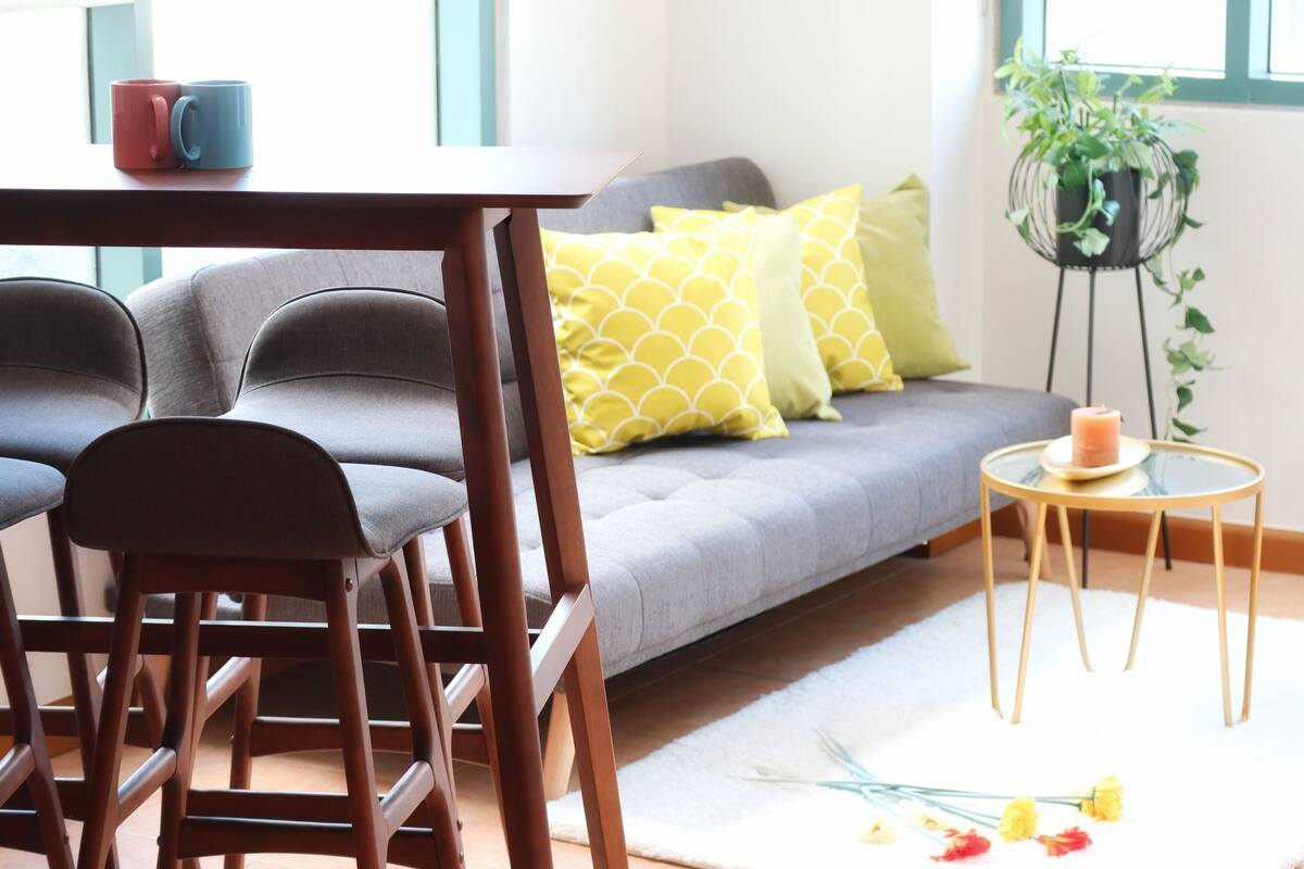 A modern living room with a grey sofa, yellow patterned pillows, a round wooden side table with a candle, and a potted plant.