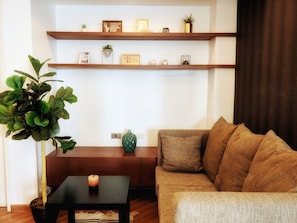 A living room with a sofa, a coffee table, and a potted plant. There are two wooden shelves above the sofa displaying various decorative items. - McKinley Park Residences (Taguig)