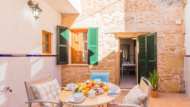 Un coin repas avec un mur en pierre, une table dressée pour le petit-déjeuner et des volets verts aux fenêtres.