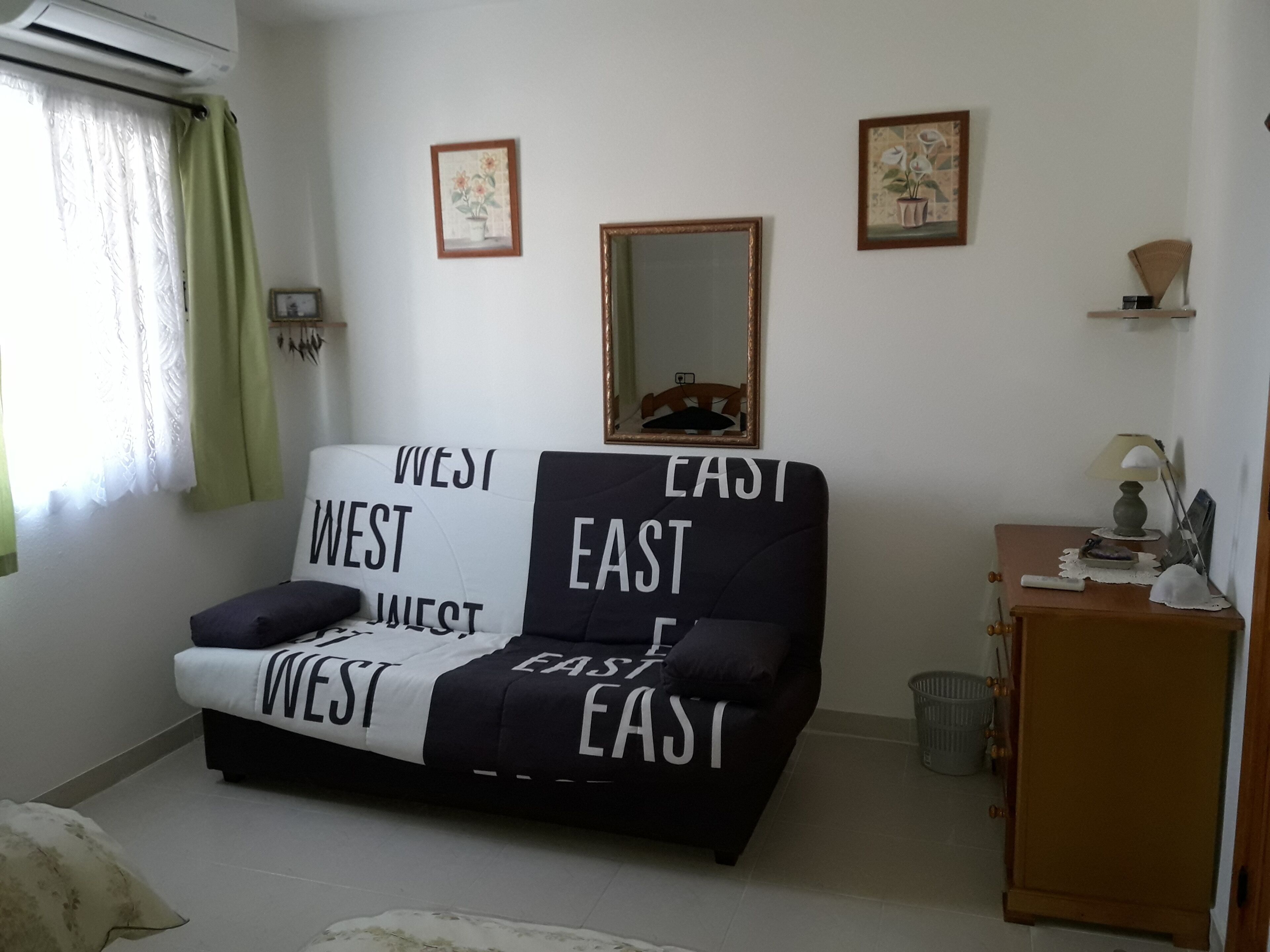Een kamer met een bank voorzien van de aanduidingen 'WEST' en 'OOST', een spiegel, een lamp en een klein kastje.