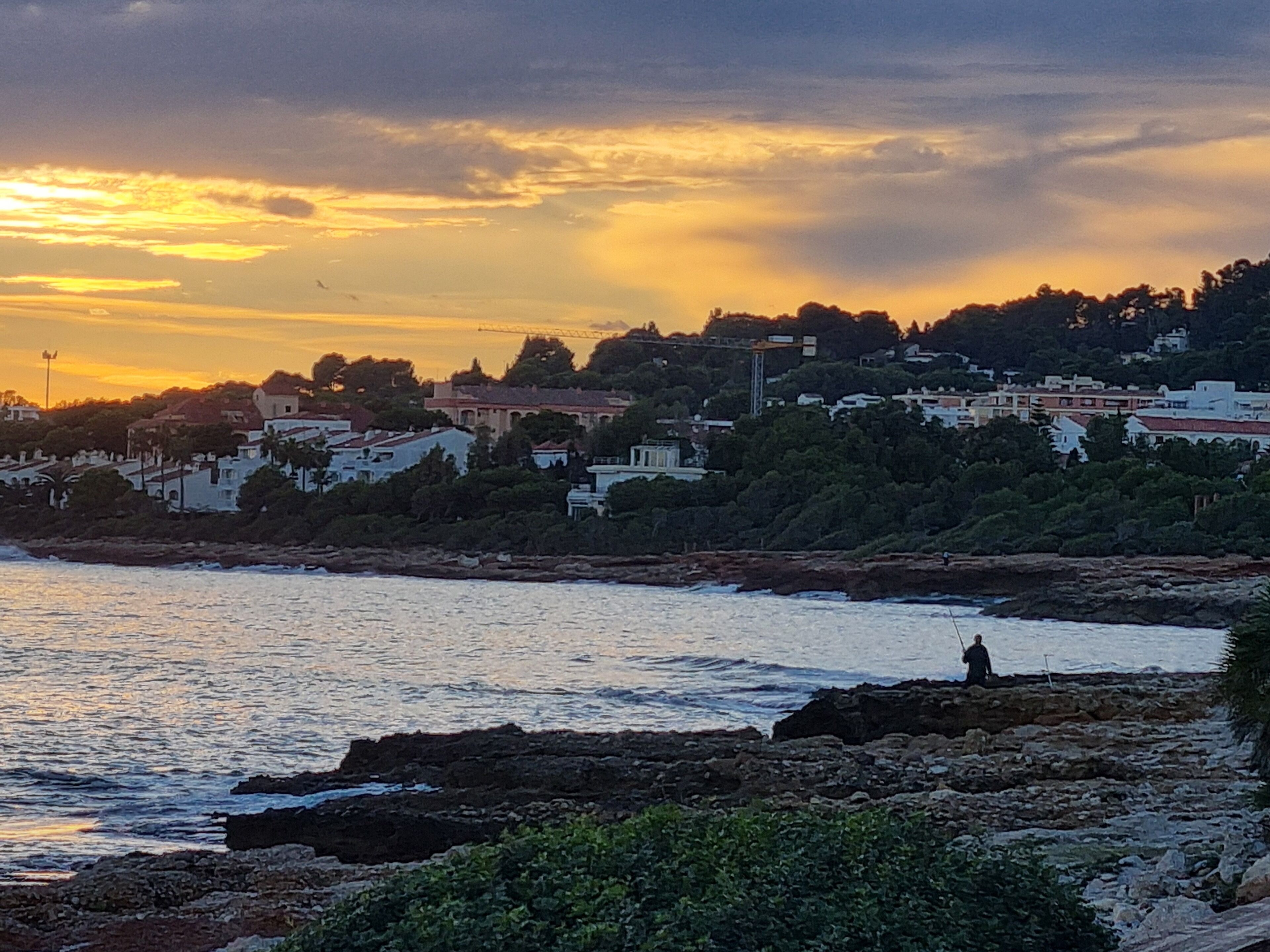 A coastal town at sunset with buildings along the hillside and a rocky shoreline.