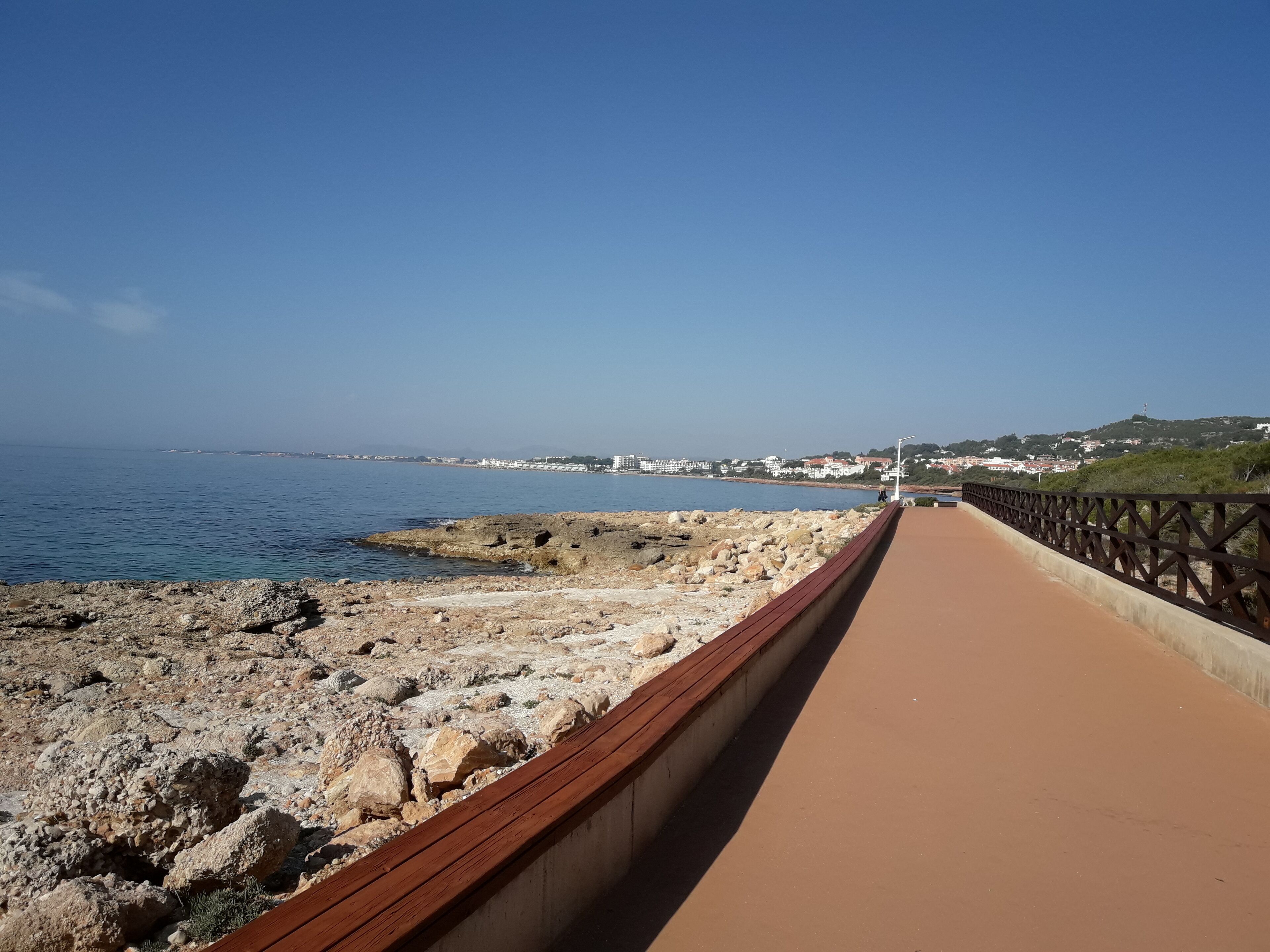 A coastal walkway with a rocky shoreline and a clear view of the sea and distant buildings.