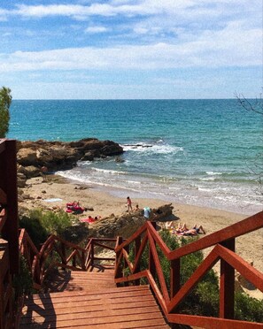 Un escalier en bois mène à une plage où l’eau est d’un bleu cristallin et où l’on trouve des affleurements rocheux.