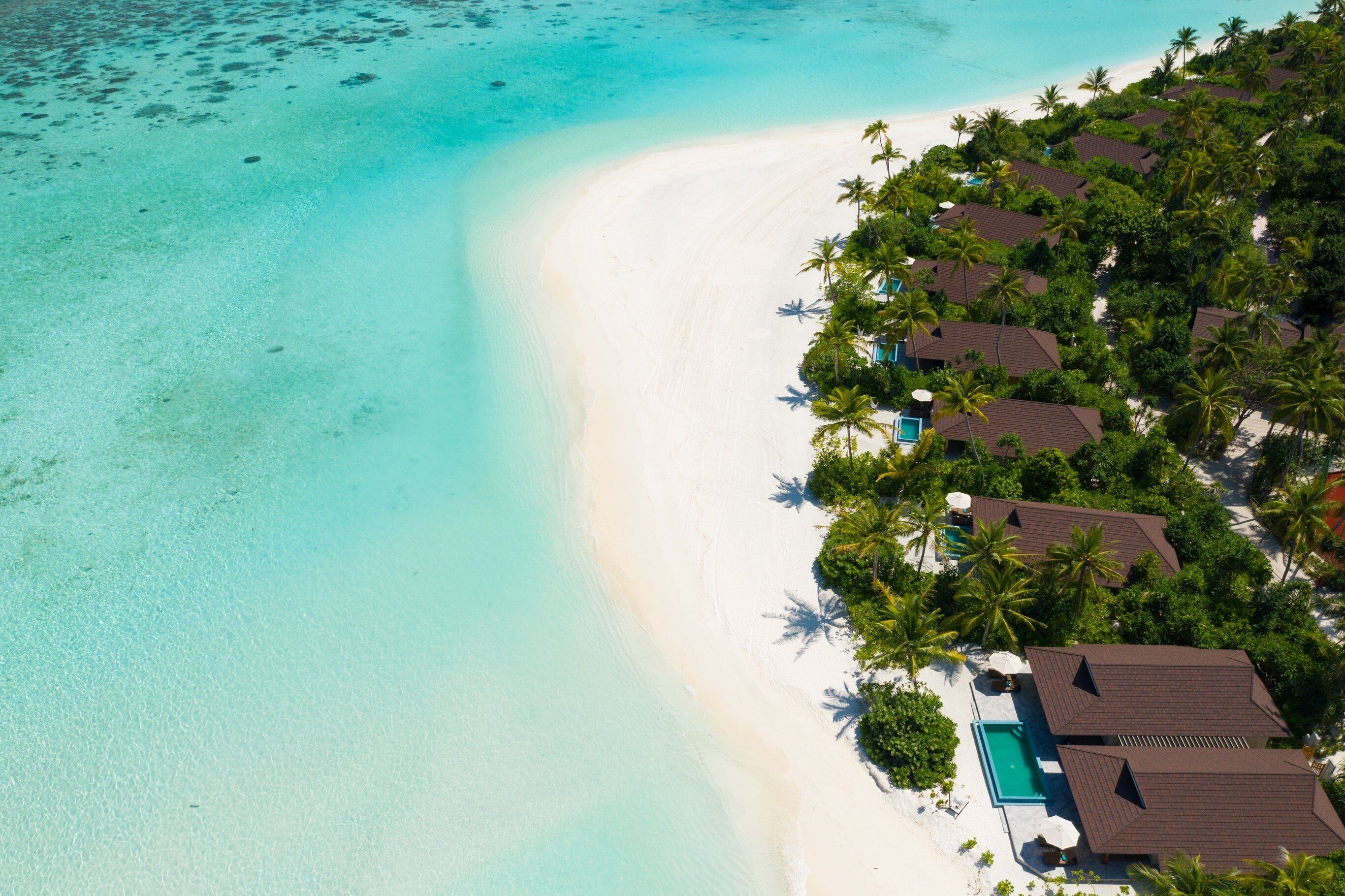 An aerial view of a beach resort with a clear turquoise sea, white sand, and a row of brown-roofed villas surrounded by palm trees.