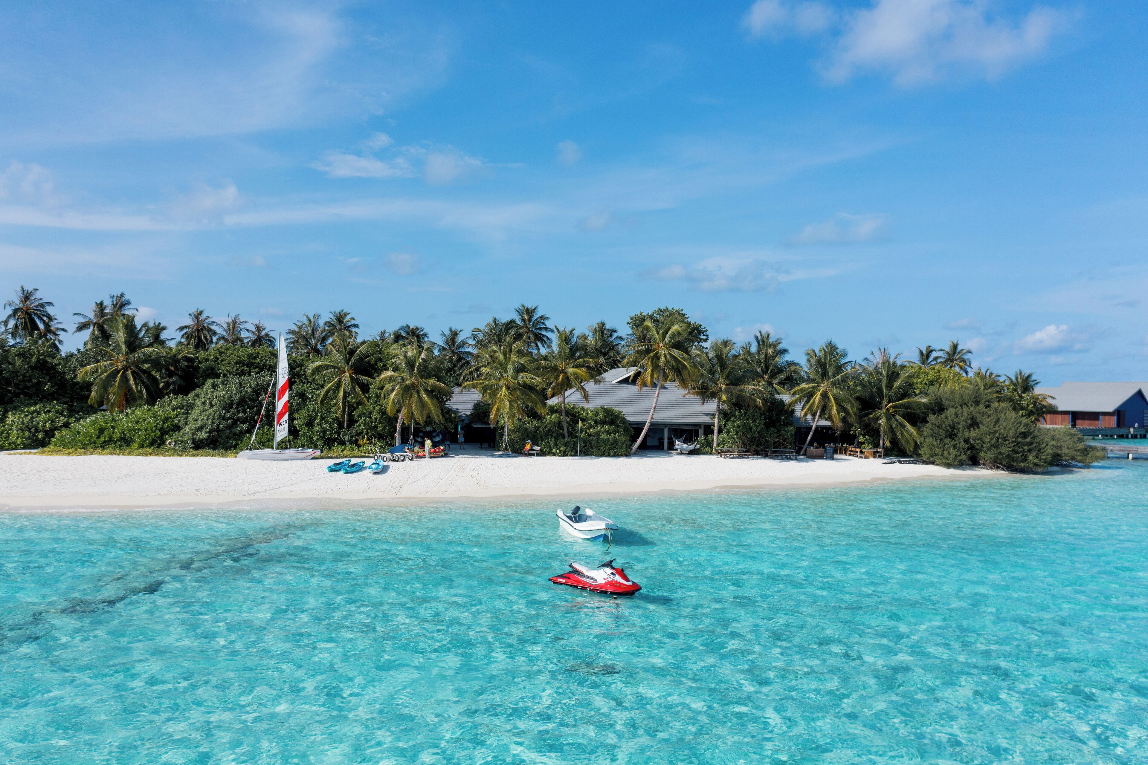 A beach with clear turquoise water, a jet ski, and a boat near a resort with palm trees and a white sandy beach.