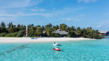 A beach with clear turquoise water, a jet ski, and a boat near a resort with palm trees and a white sandy beach.