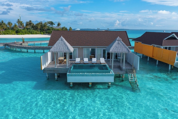 An overwater bungalow with a swimming pool, lounge chairs, and thatched umbrellas.
