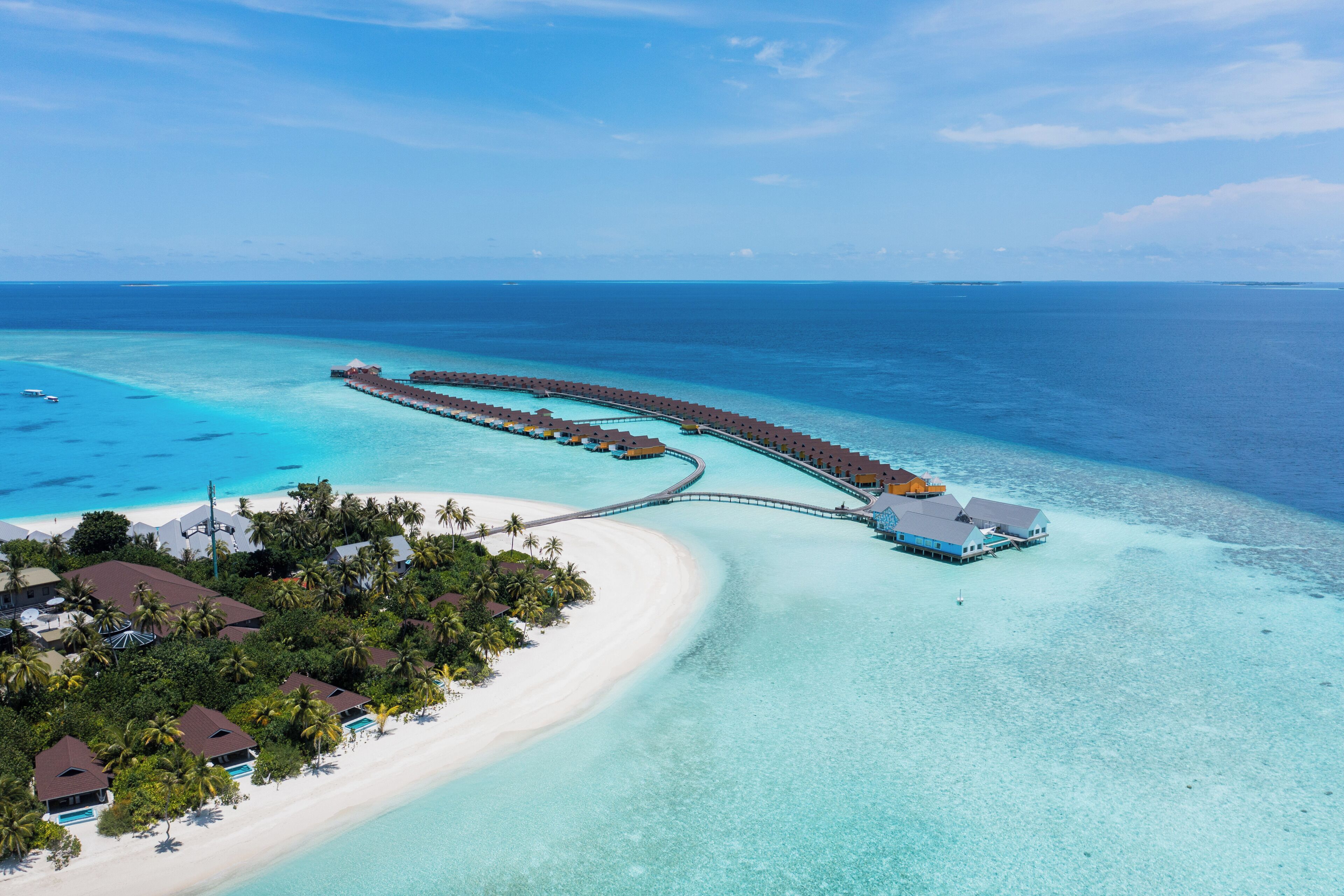 Vista aérea de un resort con un muelle largo que se adentra en aguas turquesas, rodeado de playas de arena blanca y vegetación exuberante.