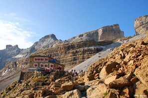 Un rifugio alpino con facciata rosso e bianco, affacciato su un pendio roccioso e con vista libera sulle pareti rocciose circostanti.