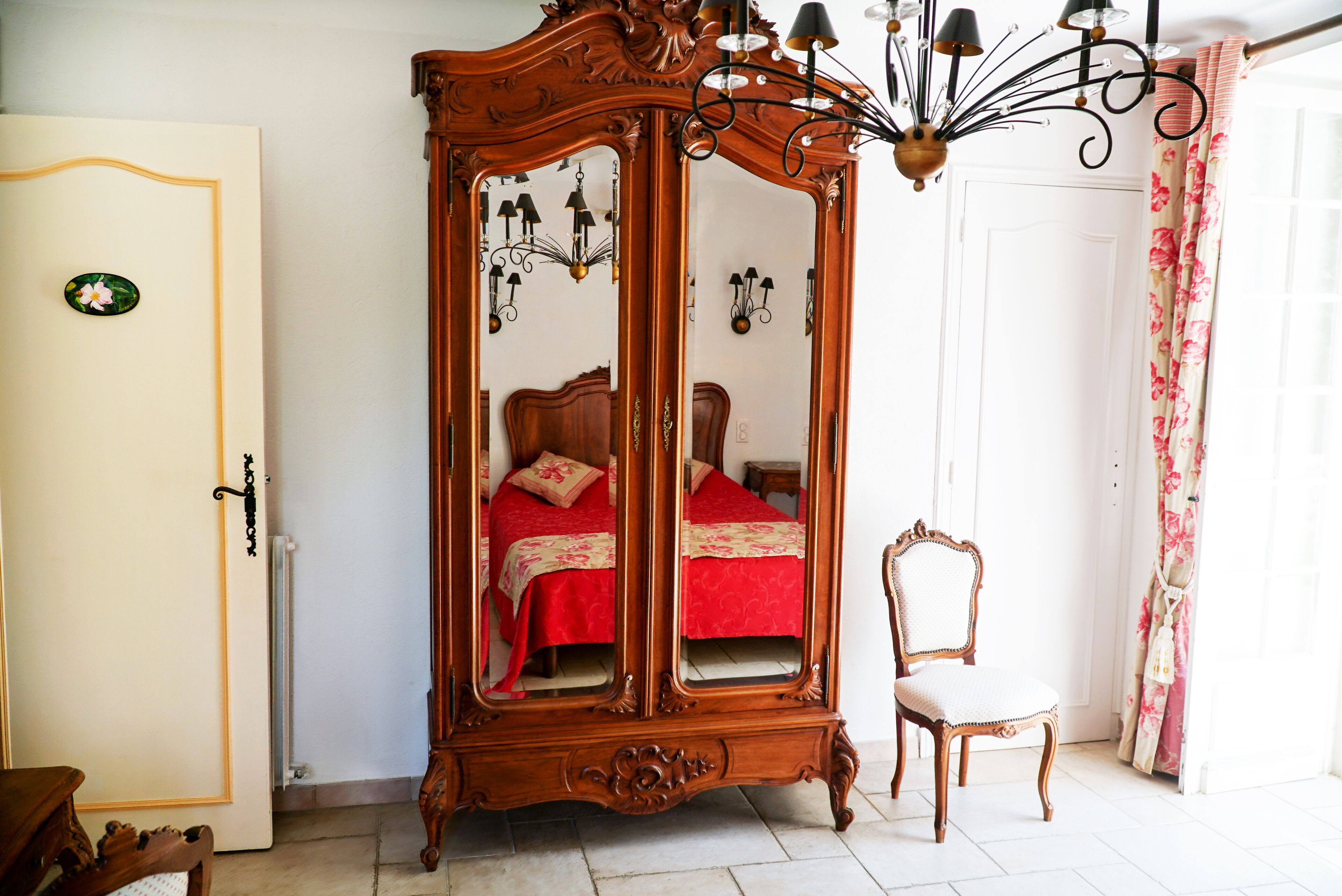 A bedroom with a large wooden armoire, a single bed with red bedding, a chair, and a chandelier.
