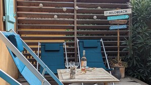 Eine Holzterrasse mit zwei blauen Liegestühlen, einem Tisch mit Flasche und Gläsern sowie Hinweisschildern.
