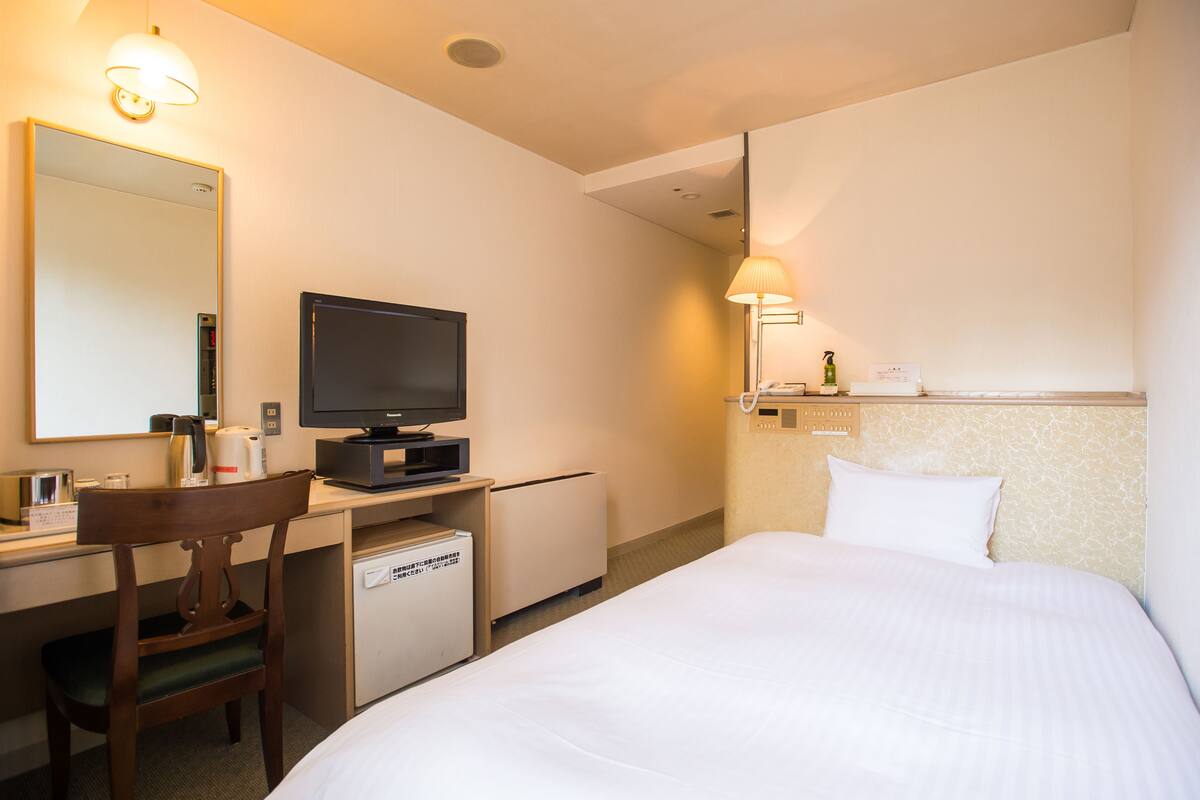 A hotel room with a bed, a desk with a television, a chair, a small refrigerator, and a bedside table with a lamp.