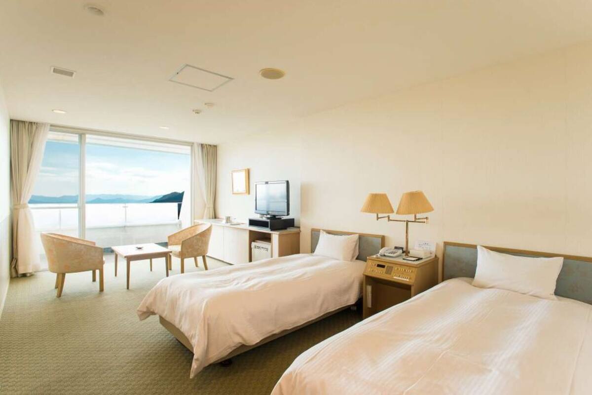 A hotel room with two beds, a desk, a chair, a TV, and a view of the sea.