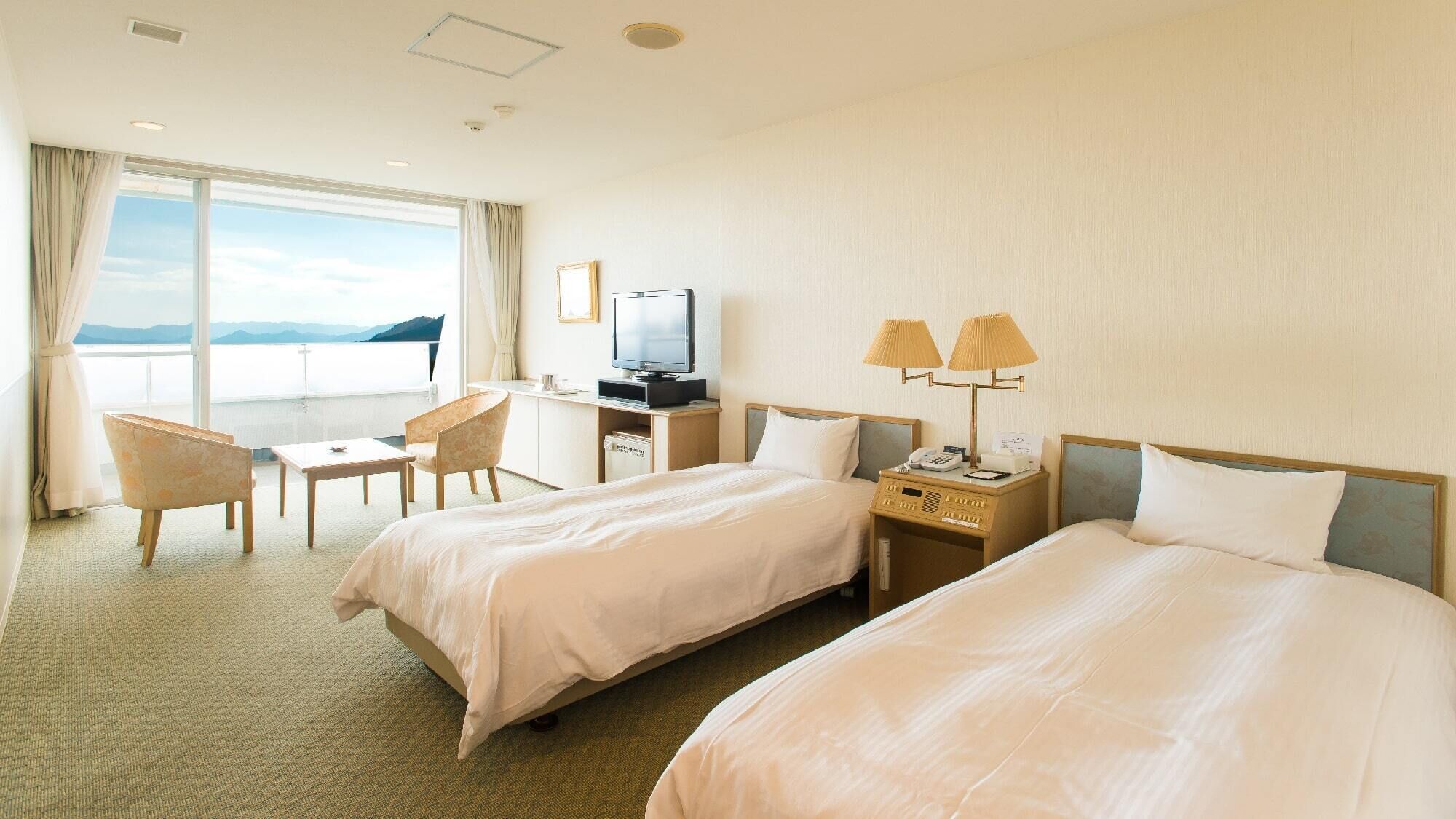 Quarto de hotel com duas camas, uma escrivaninha com televisão, uma cadeira e vista para o mar.