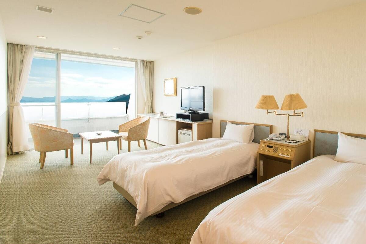 A hotel room with two beds, a desk with a TV, a chair, and a view of the sea.