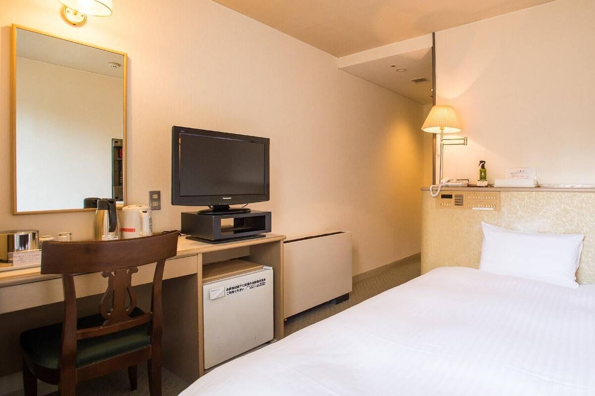 A hotel room with a bed, a desk with a television, a chair, a desk lamp, and a bedside table with a phone and a bottle.