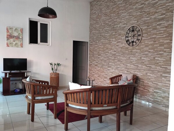 A living room with a TV, wooden chairs, a red rug, a clock, and a wall with a brick pattern.