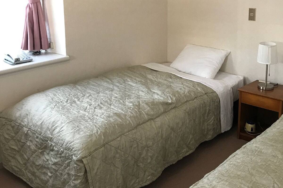A single bed with a quilted cover, a bedside table with a lamp, and a window with a curtain.