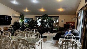 Un coin repas avec des chaises en osier et une table, un salon avec un canapé et un téléviseur, et de grandes fenêtres ornées de plantes.