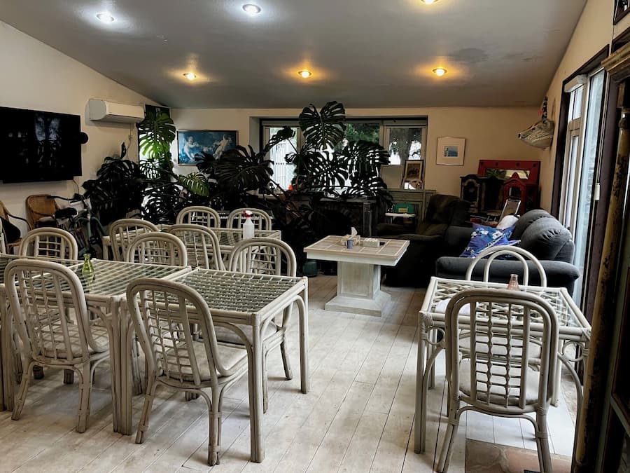 A dining area with wicker chairs and a table, a living room with a sofa and a TV, and large windows with plants.