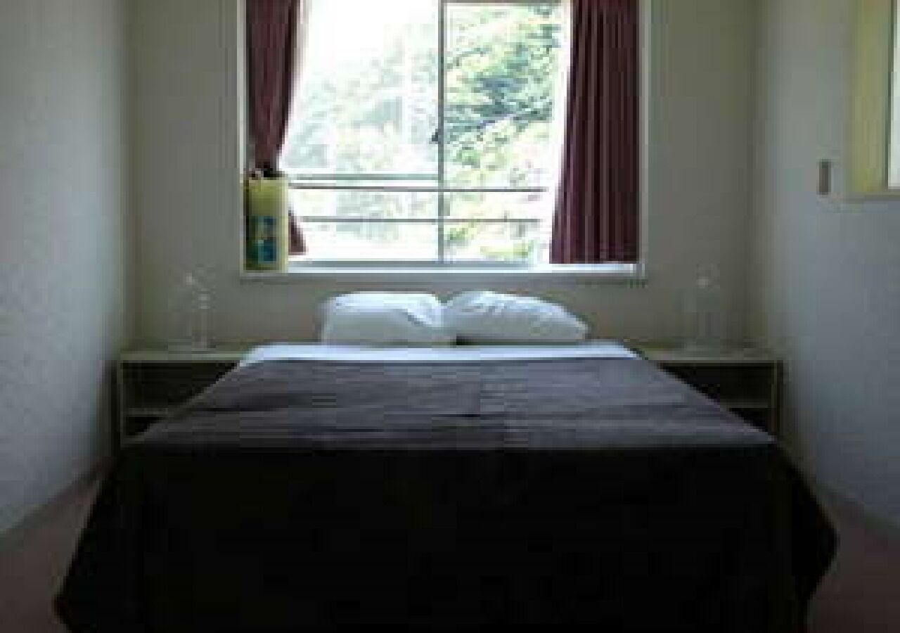 A bedroom with a bed, a window with curtains, a nightstand, and a wall-mounted lamp.