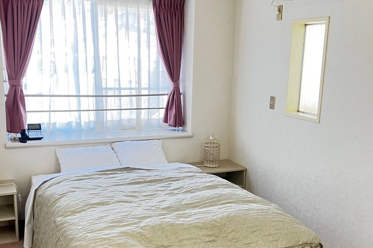 A bedroom with a bed, a bedside table, a nightstand, a window with curtains, and a wall-mounted light fixture.
