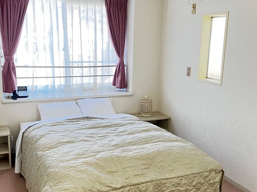 Un dormitorio con cama, mesita de noche, cortinas en la ventana y una lámpara fijada a la pared.